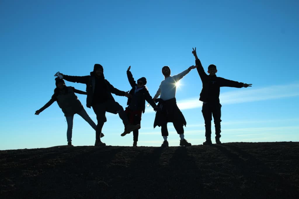 Five silhouetted figures celebrate at the summit of Acatenango Volcano by lifting their arms and legs in the air. The sun peaks behind one of the right-hand figures. The Acatenango hike difficulty depends on your fitness level and the amount of gear you need to carry.
