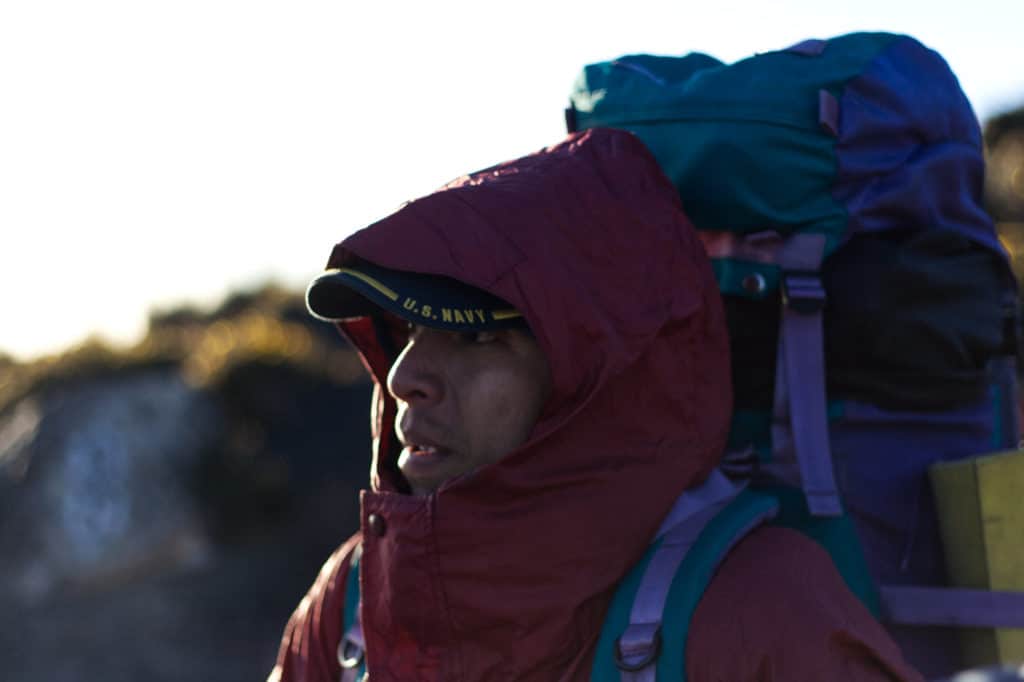 man tucked into a red coat with a hood, sunlight rimming his face and a backpack on his back.