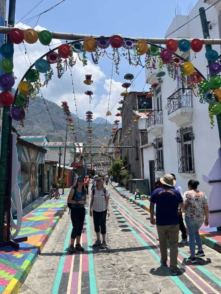 Two women stand under an arch festooned with colorful sombreros. More are hung above the whole length of the street behind them