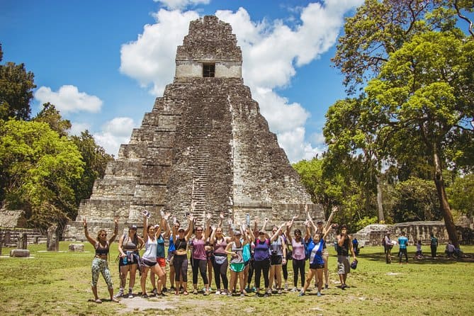 Large group of tourists stand in front of El Gran Jaguar Temple in Tikal, grinning and with their arms in the air.