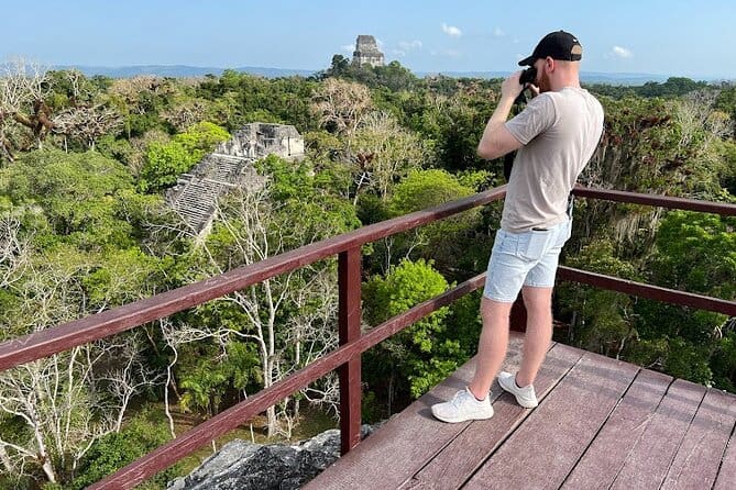 Man stands on platform high above the jungle canopy looking at the ruins below through binoculars